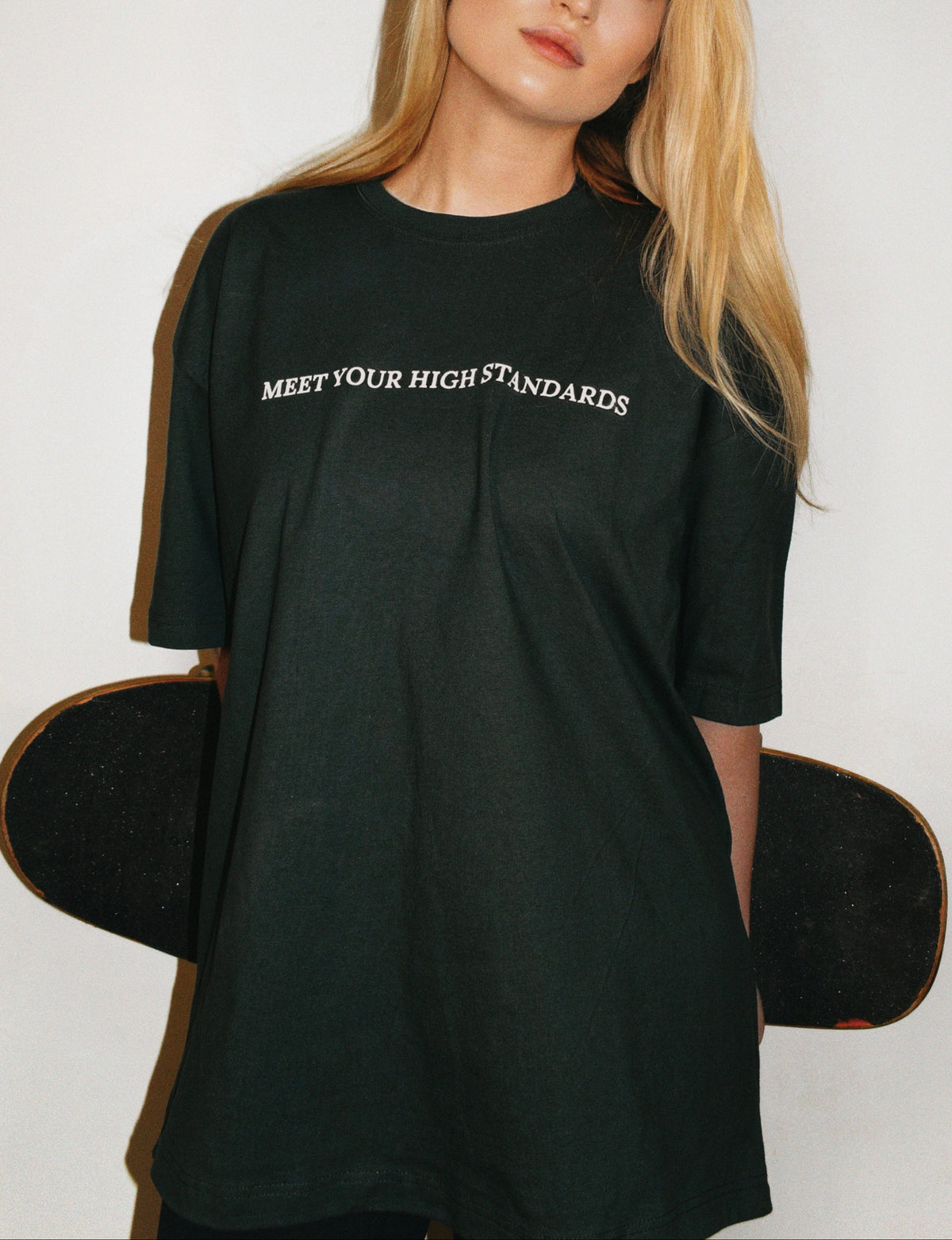 Woman wearing a green jolly cannabis / jolly athletic club t-shirt with meet your high standards text, holding a skateboard against a white background